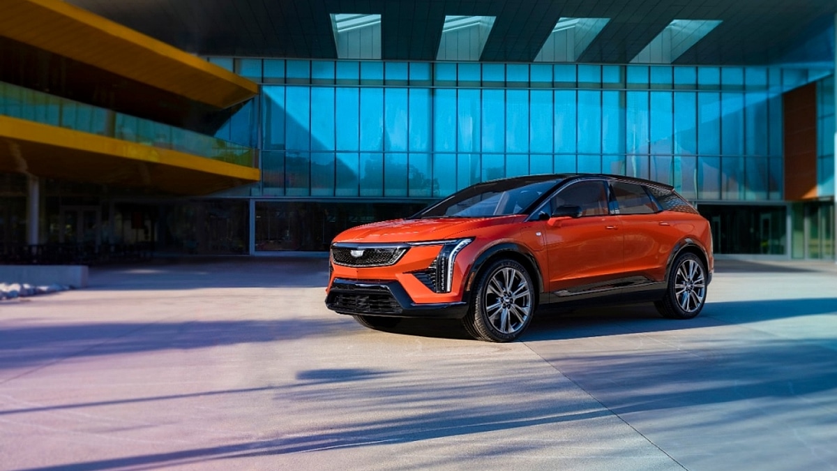 2025 Cadillac Optiq in Blaze Orange metallic posed in front of a contemporary glass and steel building with colorful architectural accents complementing the luxury electric crossovers sleek fastback coupe SUV roofline and bold proportions